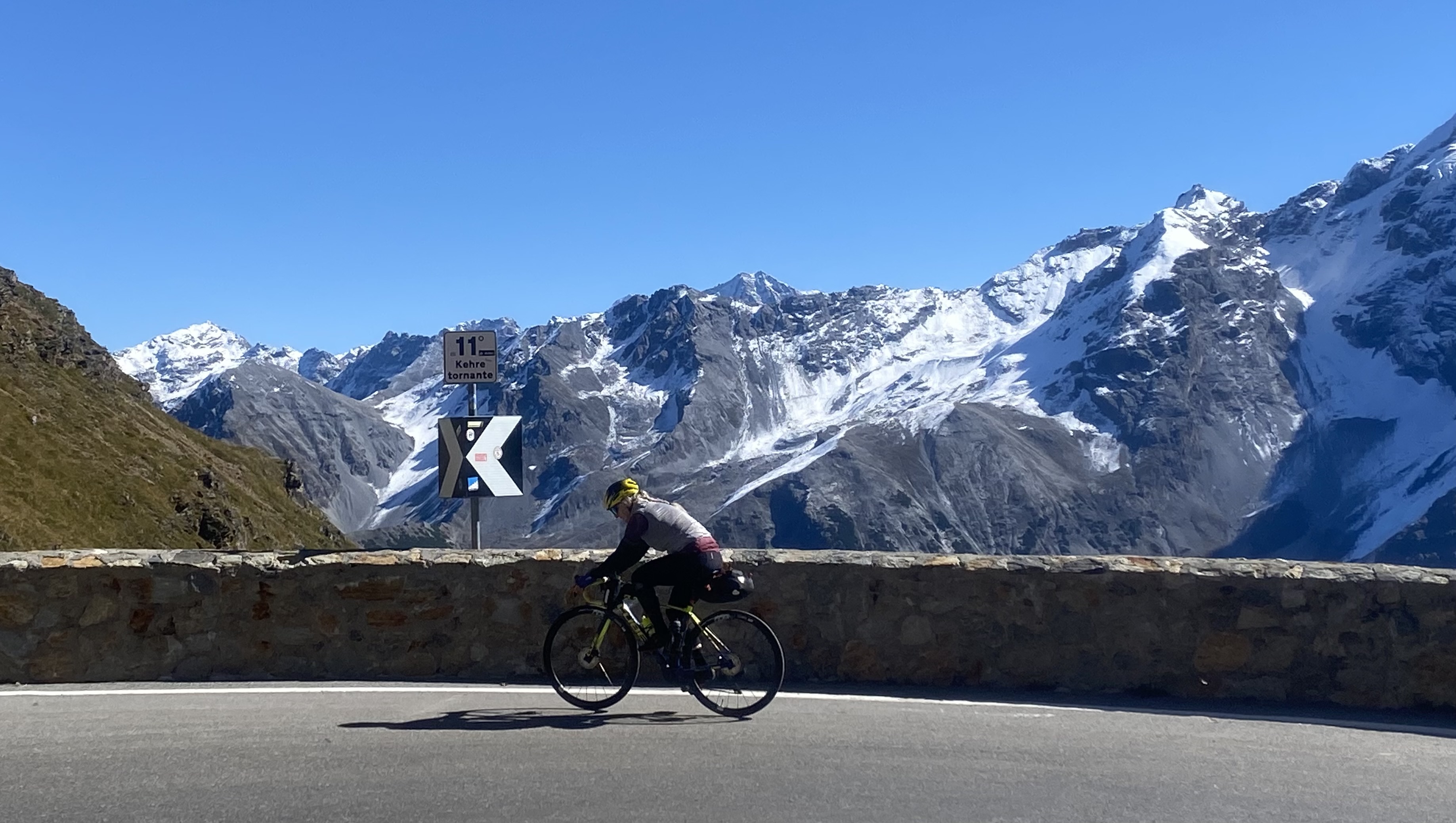 Rennradfahrerin passiert Kehre 11 der Stilfserjochstrasse (Prad-Seite); Steinmauer im Vordergrund, vergletscherte Gipfel der Ortlergruppe